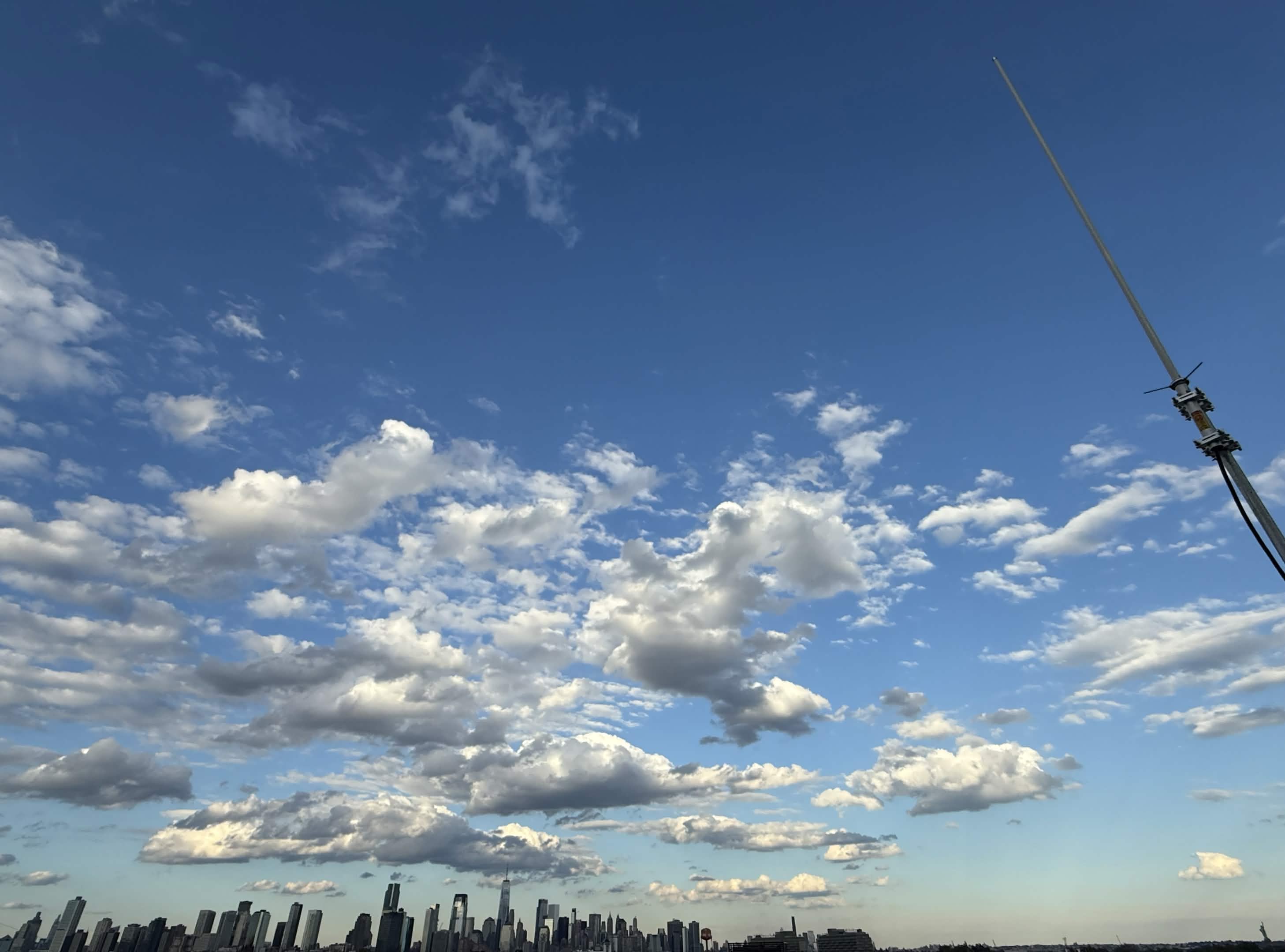 White antenna to the side of the downtown Jersey City and Manhattan skyline spanning from One Vanderbilt to the Statue of Liberty.