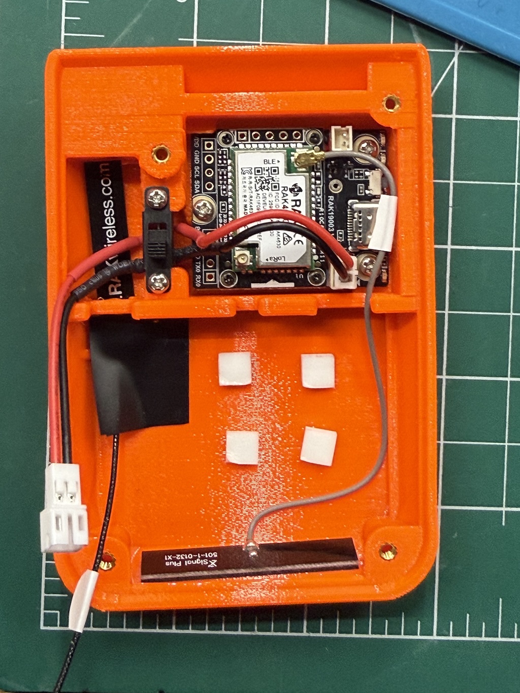 Orange plastic case with circuit board, switch, antennas inserted. Four white foam squares stuck to empty compartment.